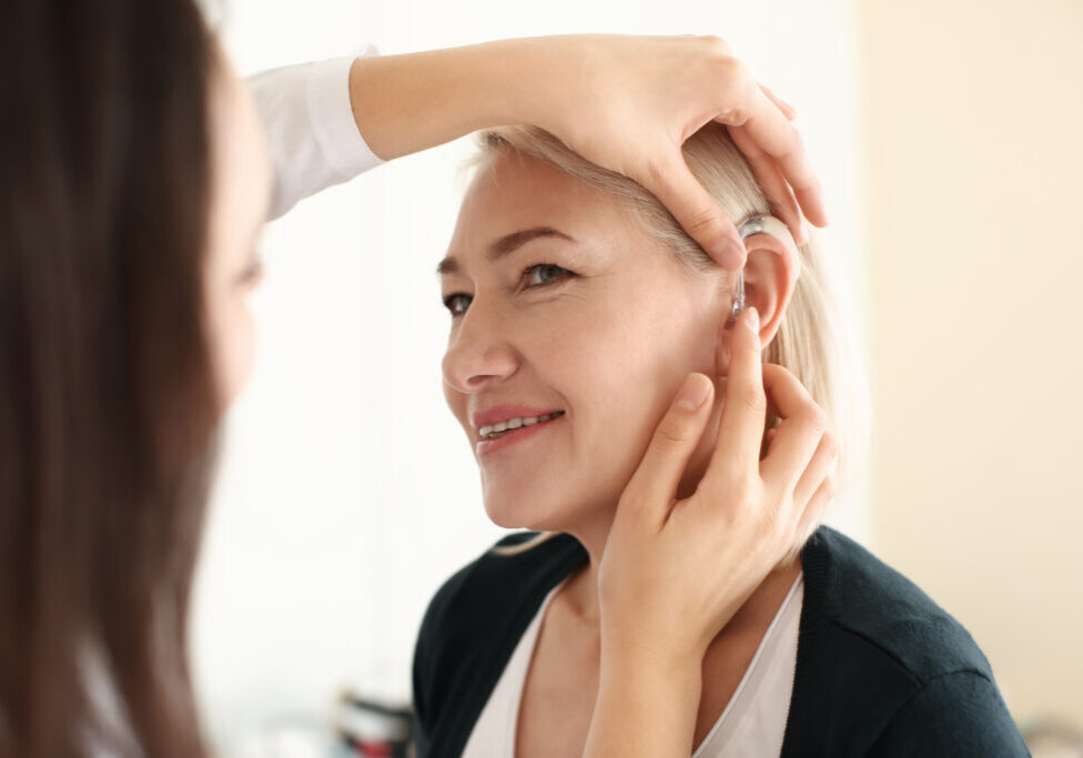 Otolaryngologist putting hearing aid in woman's ear on light background